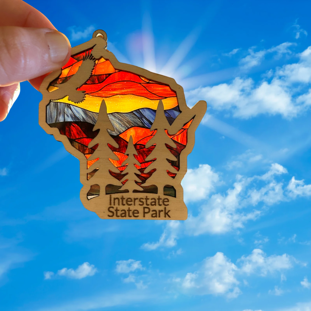 Wood Wisconsin shaped ornament with a stained glass appearance depicting an eagle soaring over trees and the text: Interstate State Park. Photo Background: ornament held up to sunlight.