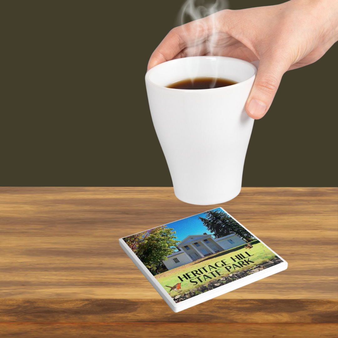 Coaster depicting a a restored historical estate house and the text “Heritage Hill State Park.” Photo background: person setting a hot beverage on the coaster.