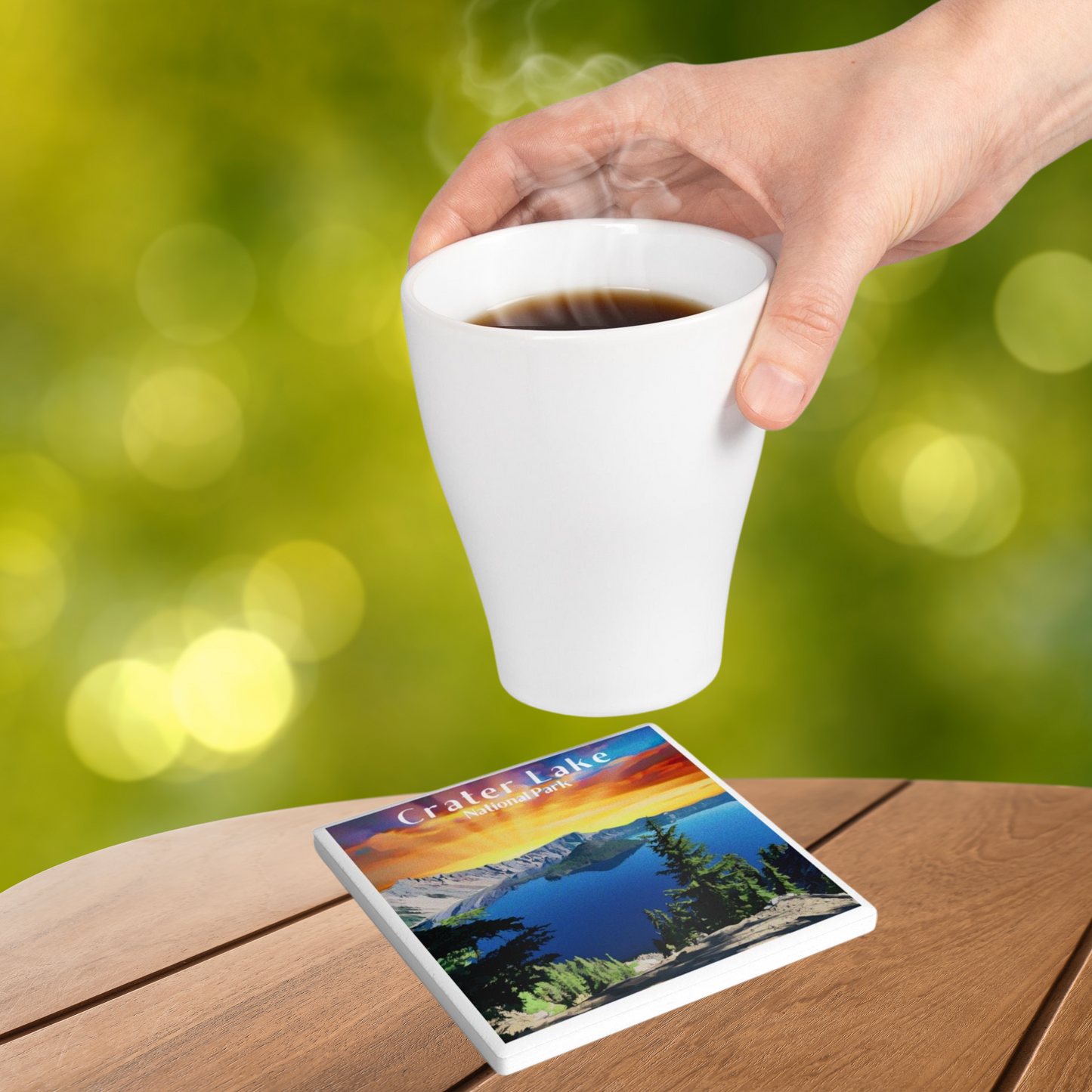 Ceramic coaster featuring a design of a vivid sunset over crater Lake and the text crater Lake national park. Photo Background: a hit beverage being set on the coaster that is sitting on a wood tabletop.