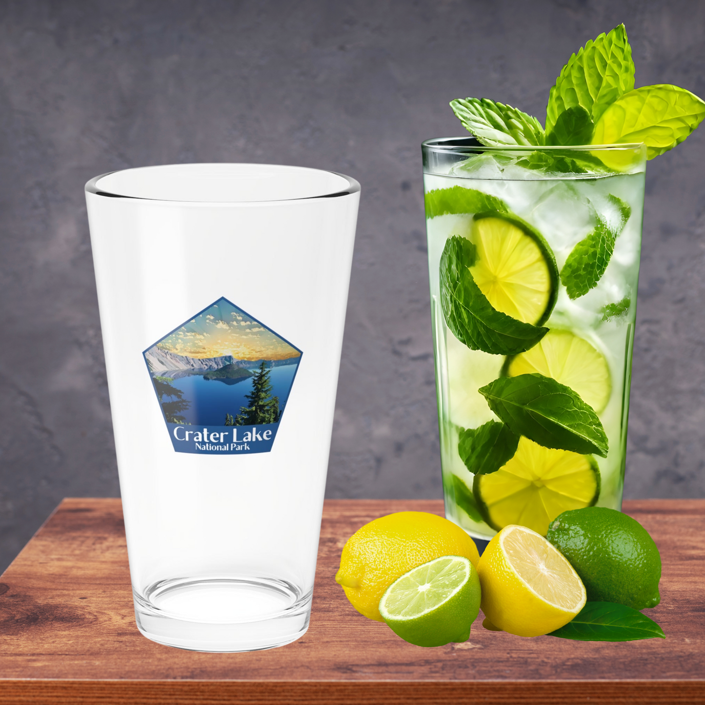 16oz pint glass sitting on a wood counter with lemons, limes, and a glass full of water and lime wedges with mint leaves. Pint glass features a pentagon shaped design of a sunrise over Crater Lake and the Text Crater Lake National Park.