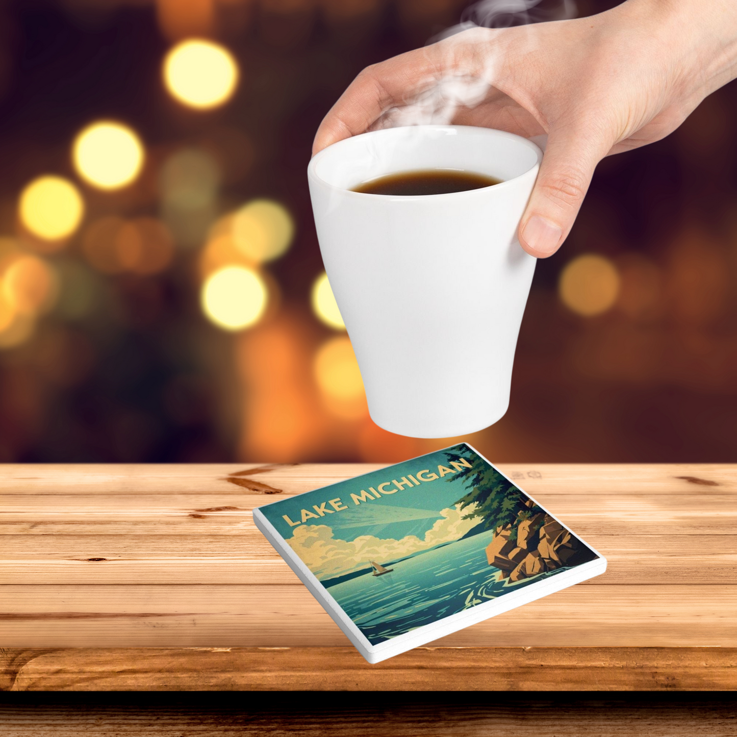 Ceramic coaster design featuring a rocky shoreline with trees and a vast lake with a sail boat, blue sky with white clouds and the text Lake Michigan. Photo Background: a hot beverage being set on a coaster that is set on a rustic wood table top.