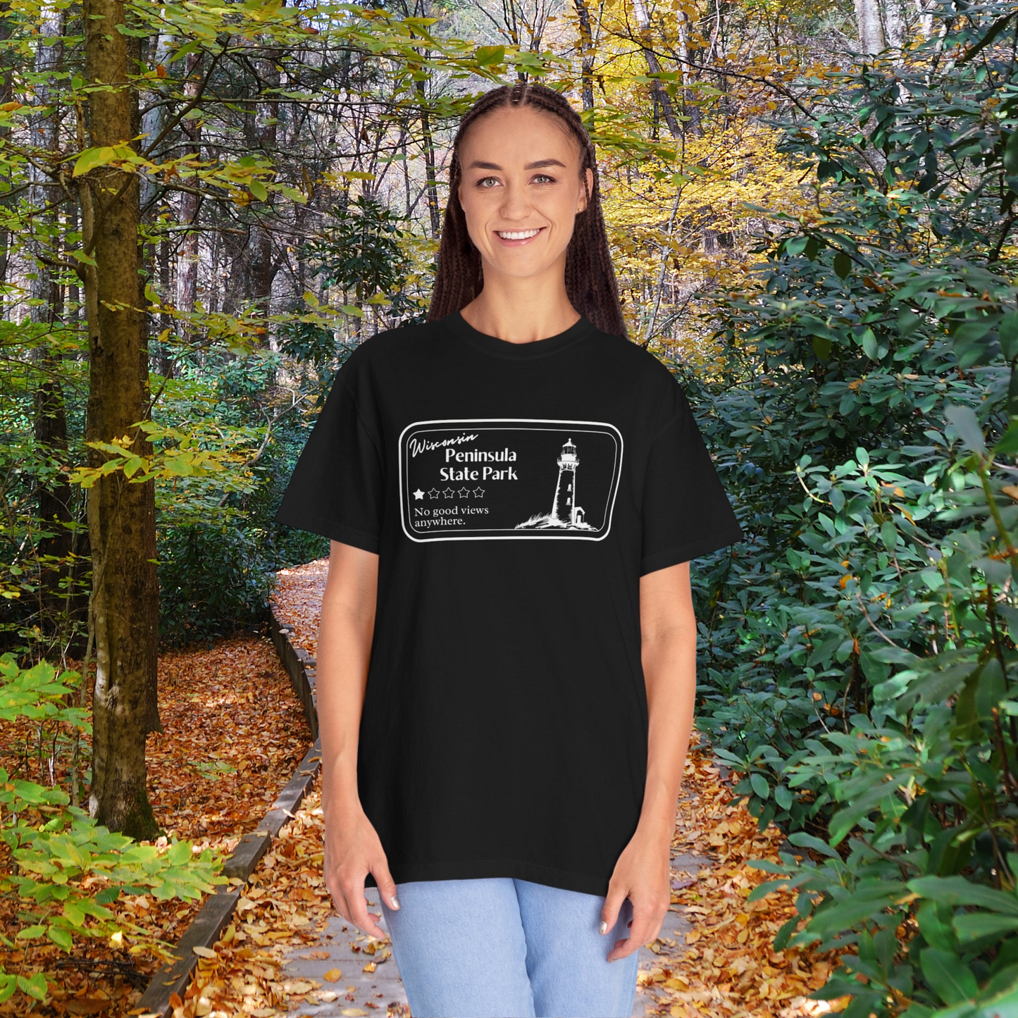 Female model wearing a Black T-Shirt with 1 star review design of Peninsula State Park. Features a line art image of a lighthouse, 1 star graphic, and the text: “Wisconsin Peninsula State Park, No good views anywhere. Model is standing on a trail in the woods.