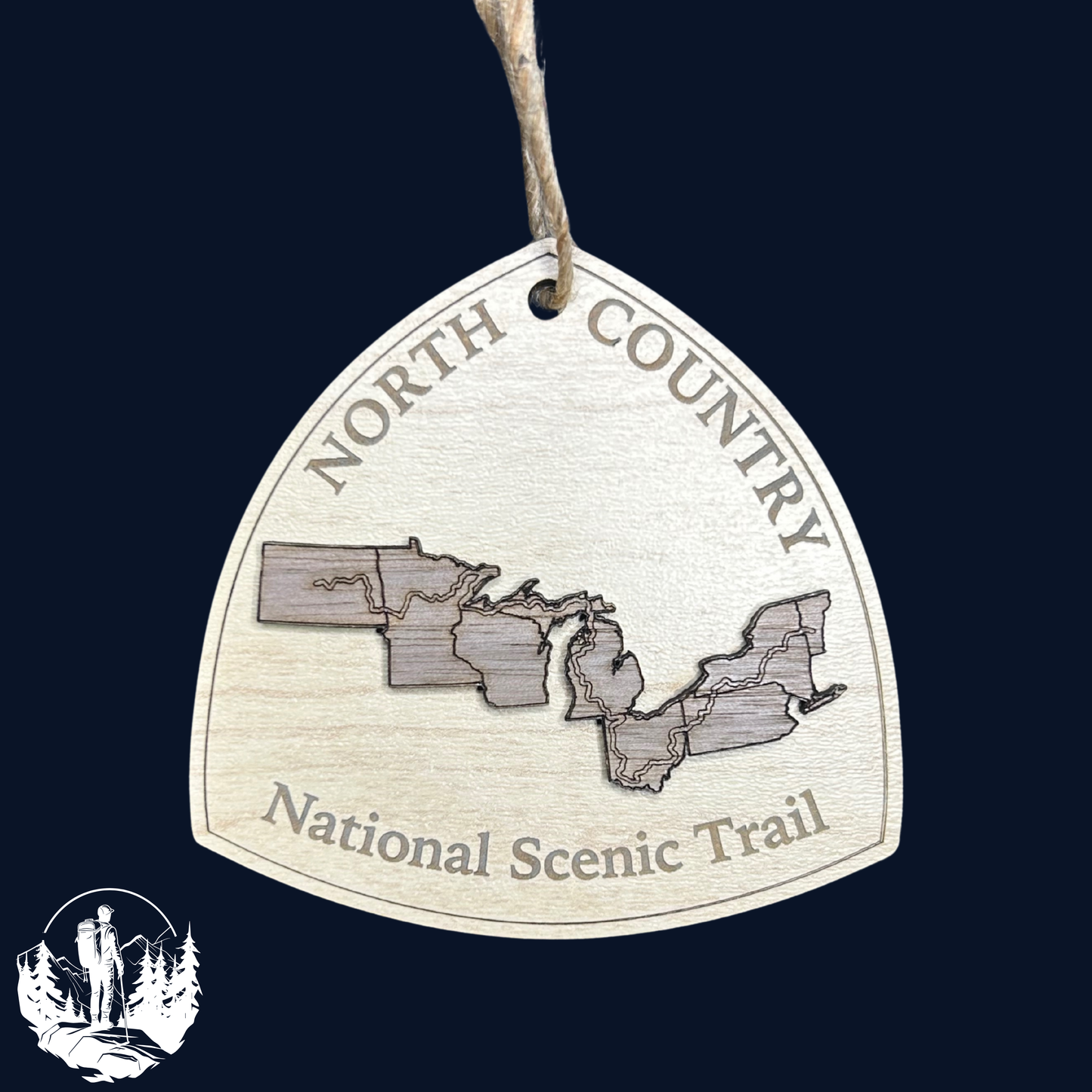 Wood ornament in the shape of the national trail's icon, with a walnut North Dakota, Minnesota, Wisconsin, Michigan, Ohio, Pennsylvania, New York, and Vermont Shaped center piece depicting the North Country Trail Route. Ornament contains the text "North Country National Scenic Trail". Dark color background.