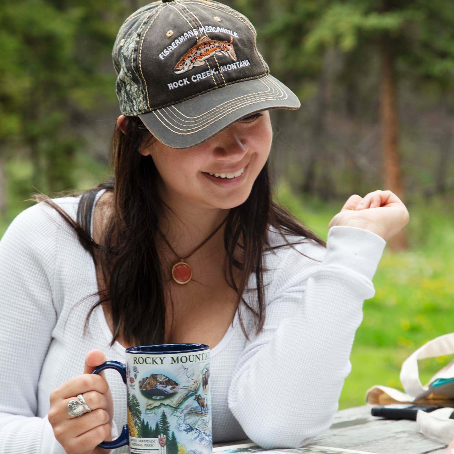 A woman outdoors wearing a dark camouflage baseball cap with 'Rocky Mountain Fishing Recreation' and 'Rocky Mountains' printed on it, along with a white long-sleeve top and holding a printed ceramic mug.