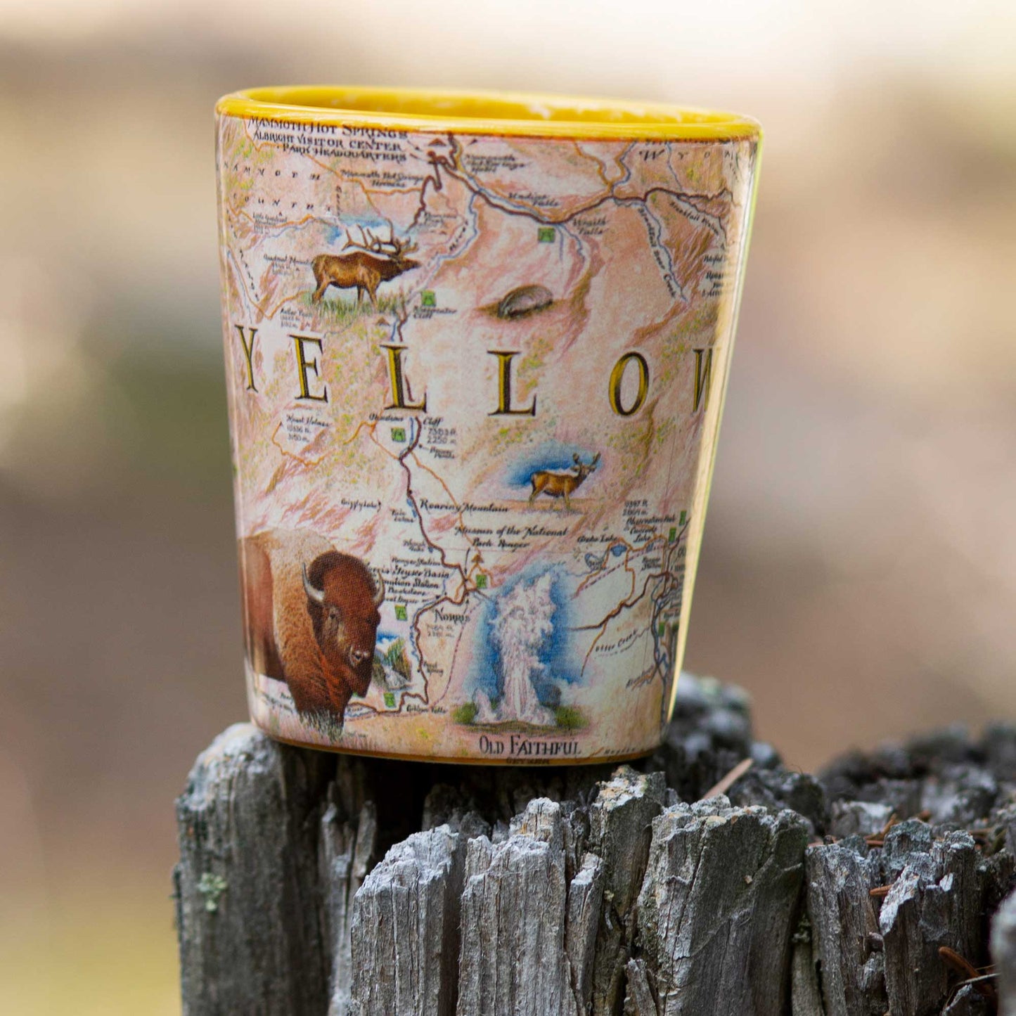 A novelty shot glass featuring a vintage-style map of Yellowstone National Park with illustrations of wildlife, resting on a weathered tree stump outdoors.