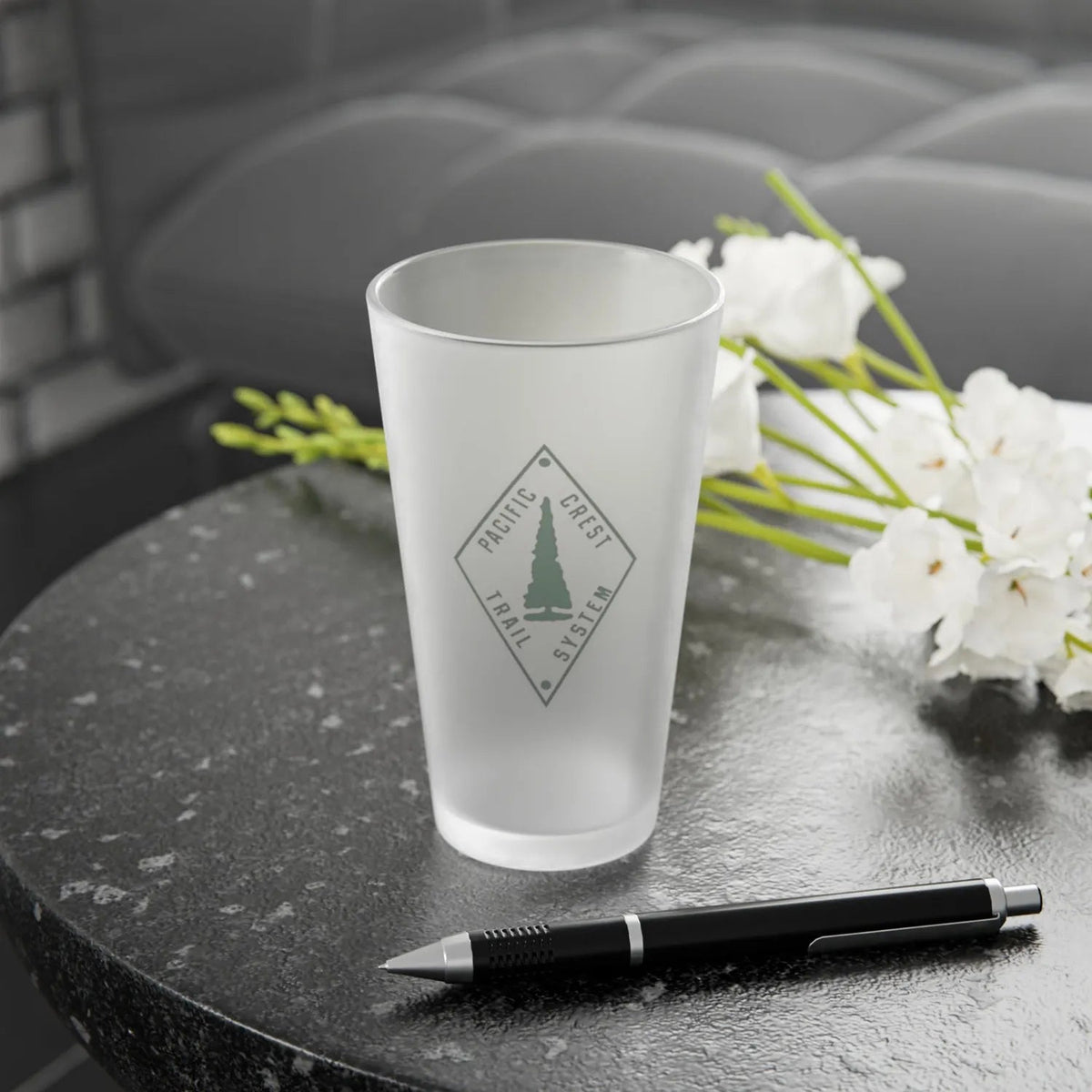 Frosted glass with the Pacific Crest Trail logo on a dark surface with white flowers and a pen.