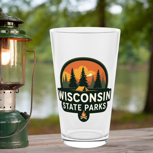 16 oz glass pint featuring a colorful design of a sunset over a campsite surrounded by pine trees and the text Wisconsin State parks.  Photo background: glass sitting on picnic table with a lantern near a lake.