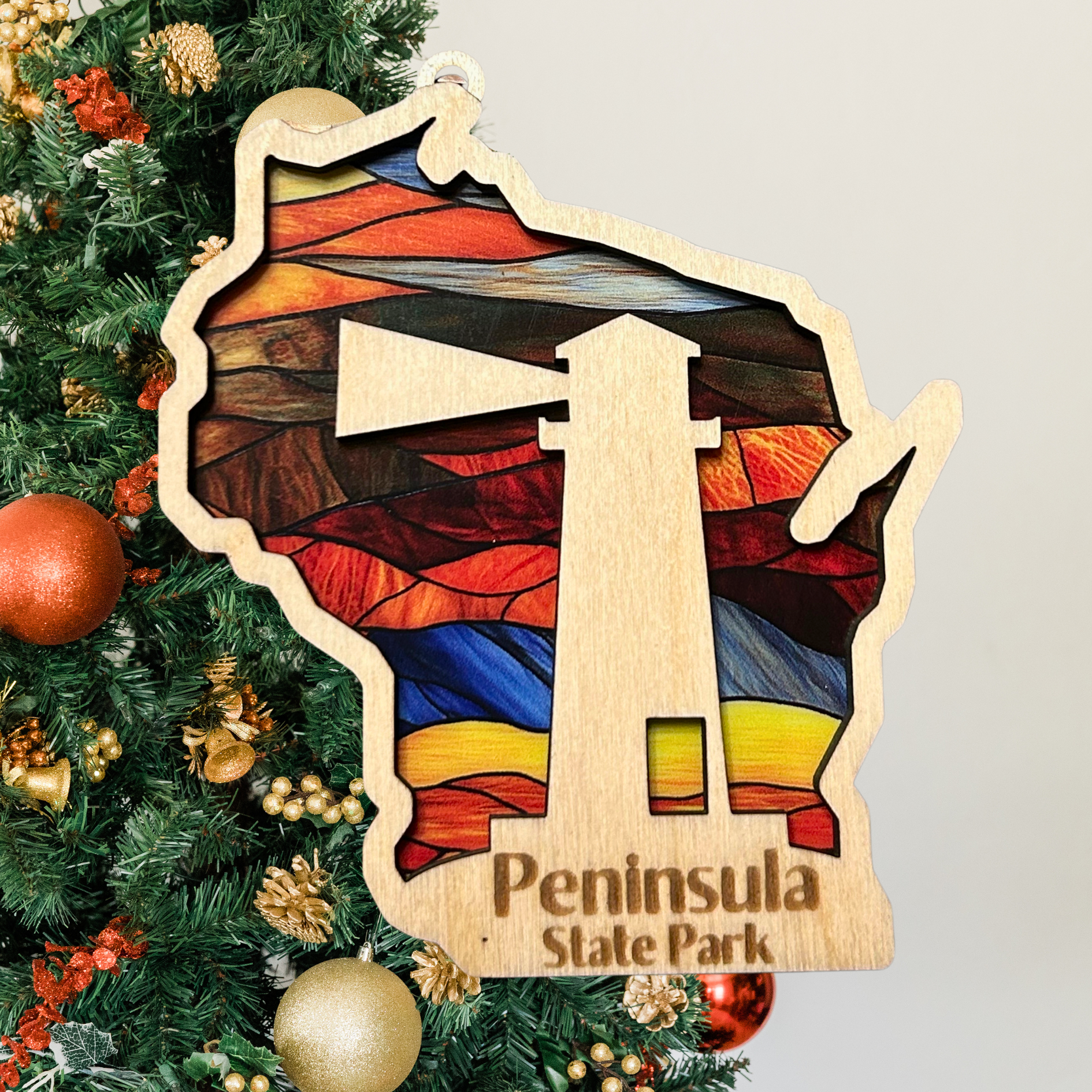 Wood Wisconsin Shaped ornament with a stained glass look and a lighthouse.  Includes the text: Peninsula State Park. Photo Background: Christmas tree.