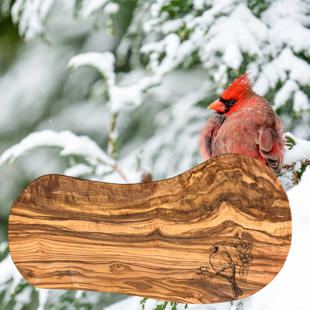 Olive wood serving board with an engraving of a cardinal sitting on a flowering branch.  Photo background: Cardinal amongst snow covered trees.