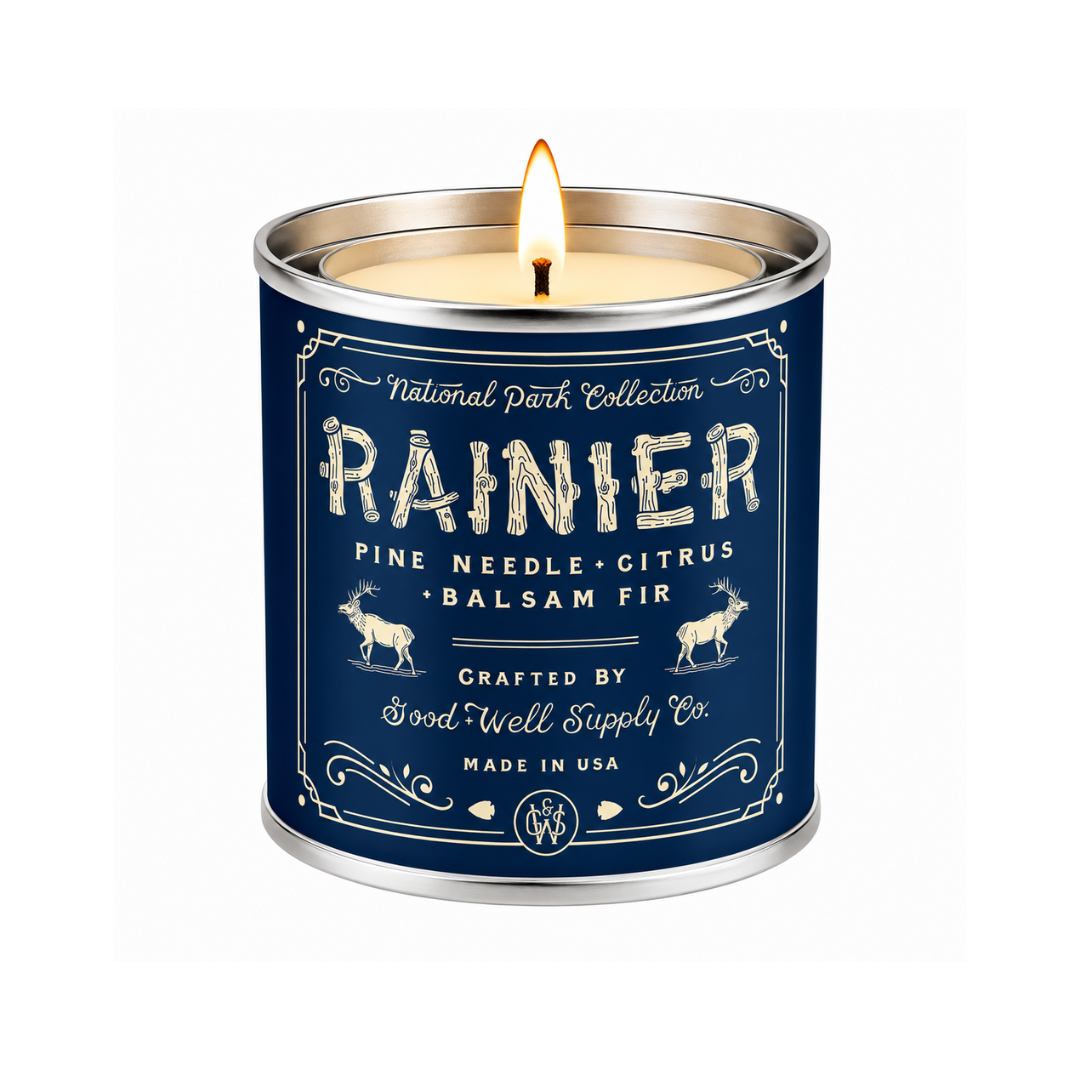 A lit tin candle from the National Park Collection, specifically Rainier, with a navy blue label and silver metal container, shown against a plain white background.