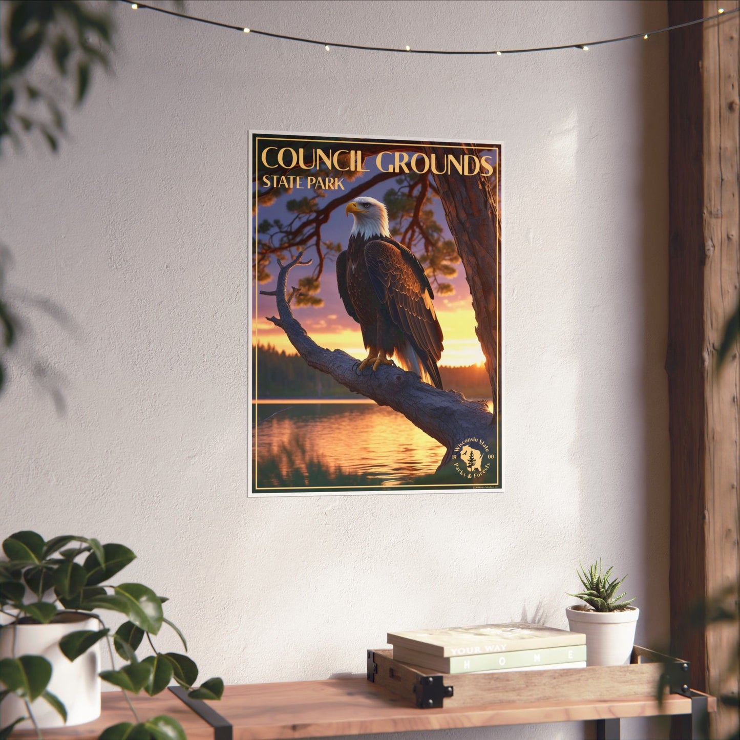 Poster depicting a majestic bald eagle sitting on a pine branch over looking a pristine lake at sunset, and the text Council Grounds State Park. Photo background: poster hanging a wall above a wood shelf with plants and books.