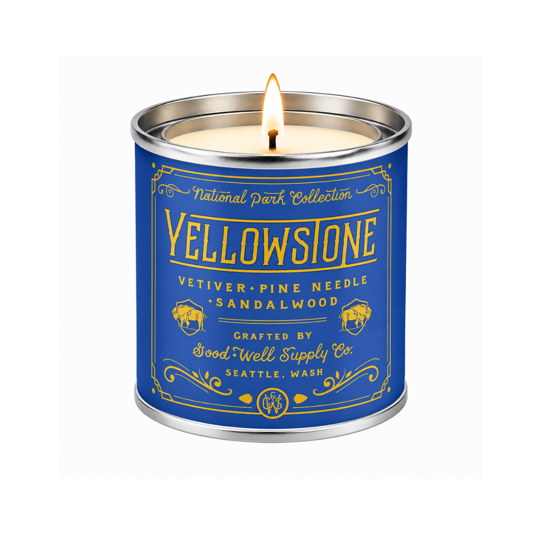 A lit scented candle in a cylindrical metal tin with a blue label and gold lettering. The label reads 'Yellowstone' and 'National Park Collection' with 'Vetiver-Pine Needle - Sandalwood' and 'Crafted by Good Well Supply Co.