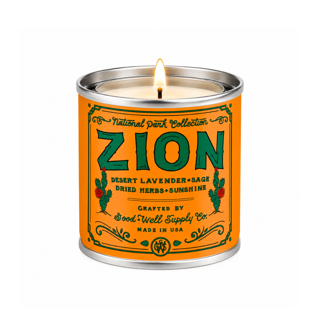 A lit scented candle in a cylindrical metal tin with a bright orange label reading “ZION” and “National Park Collection,” shown on a plain white background.