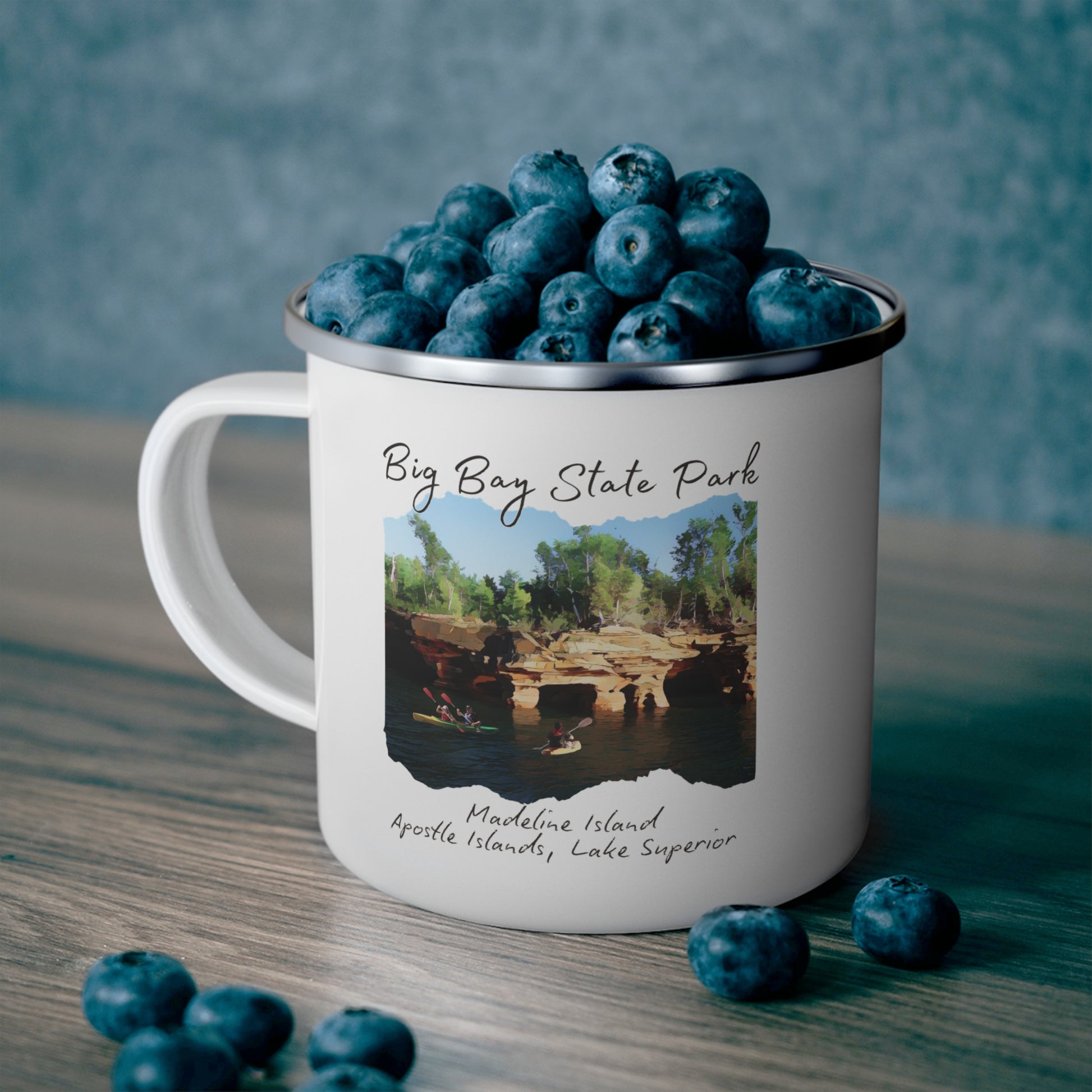 White enamel mug with picture depicting kayakers in front of sea caves and the text “Big Bay State Park - Madeline Island, Apostle Islands, Lake Superior.  Photo background: Mug full of blueberries sitting on counter.