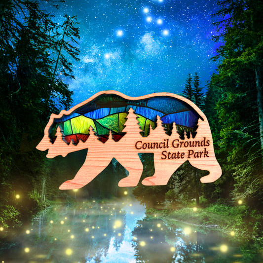 Handmade cedar bear shaped magnet with a forest scene and stained glass appearance and the text: Council Grounds State Park.  Photo background:  Starry night in a forest with firefly’s flying above a calm creek.