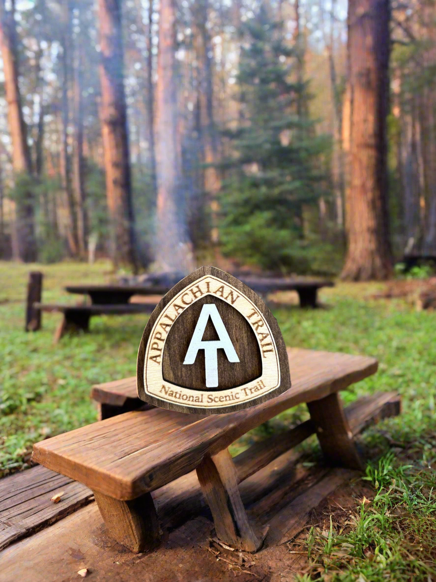 National scenic trail shaped magnet with Appalachian Trail National Scenic Trail, and white AT text symbol.  Background is primitive campsite with picnic tables, green grass, and trees.