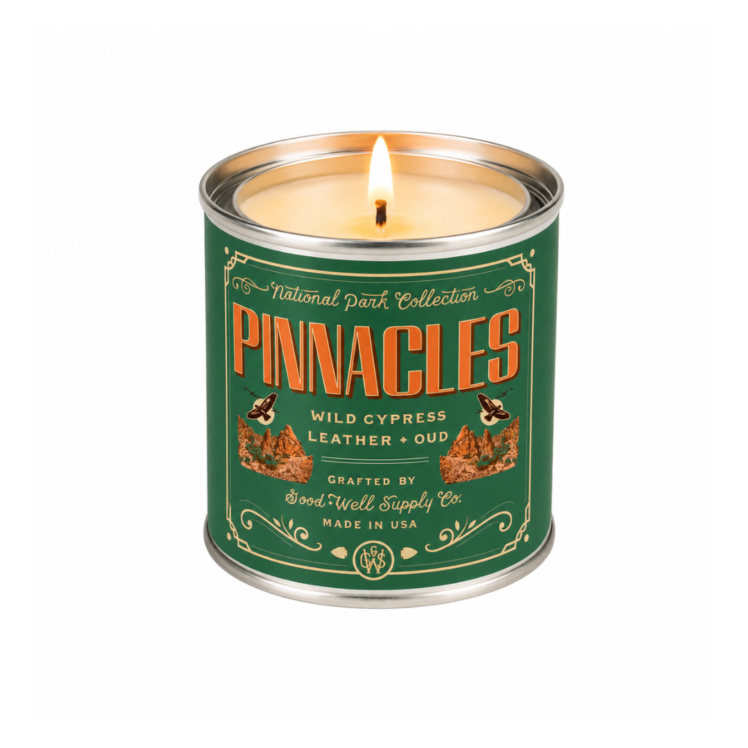 A lit metal tin candle from the National Park Collection. The candle is labeled 'Pinnacles' with 'Wild Cypress + Leather + Oud' and features a dark green label with orange lettering and illustrations of cypress trees and bees, set against a plain white background.