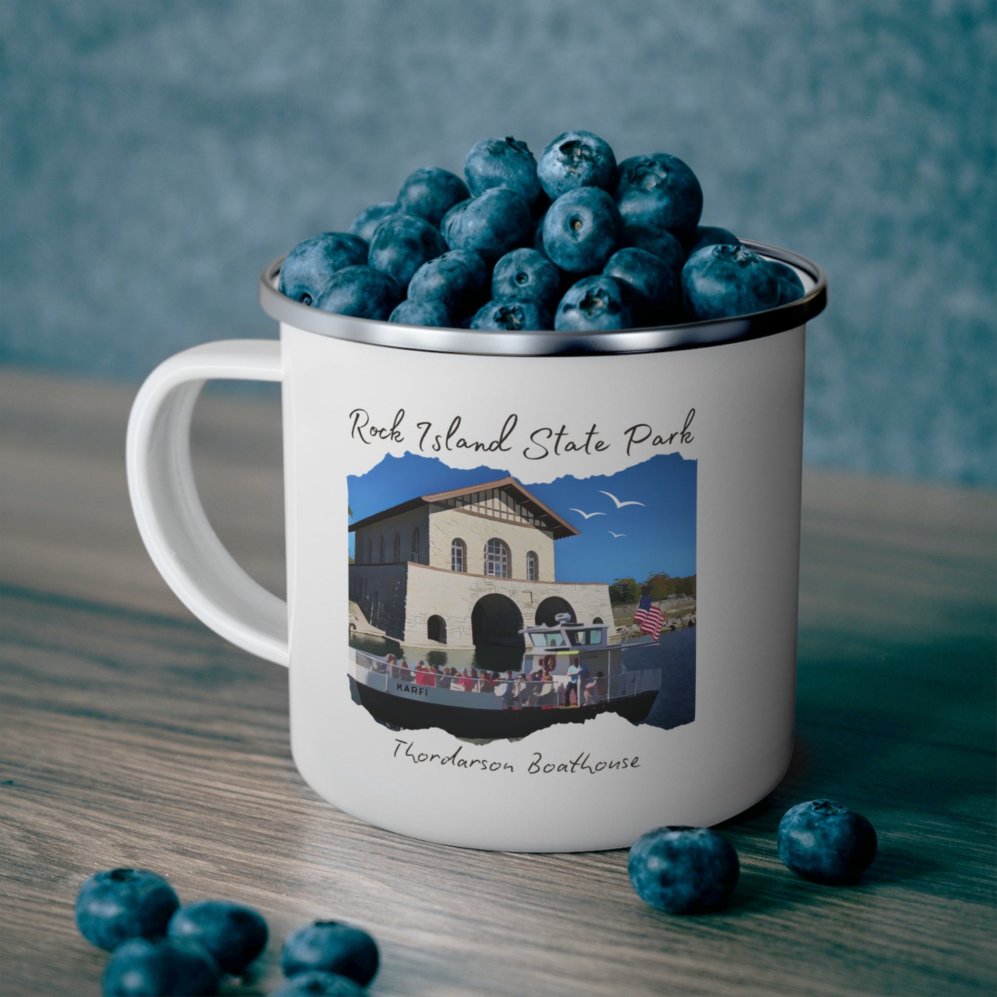 White enamel campfire mug depicting a scene of a ferry boat pulling up to the stone boat house on Rock Island.  Photo background: mug full of blueberries sitting on a counter.