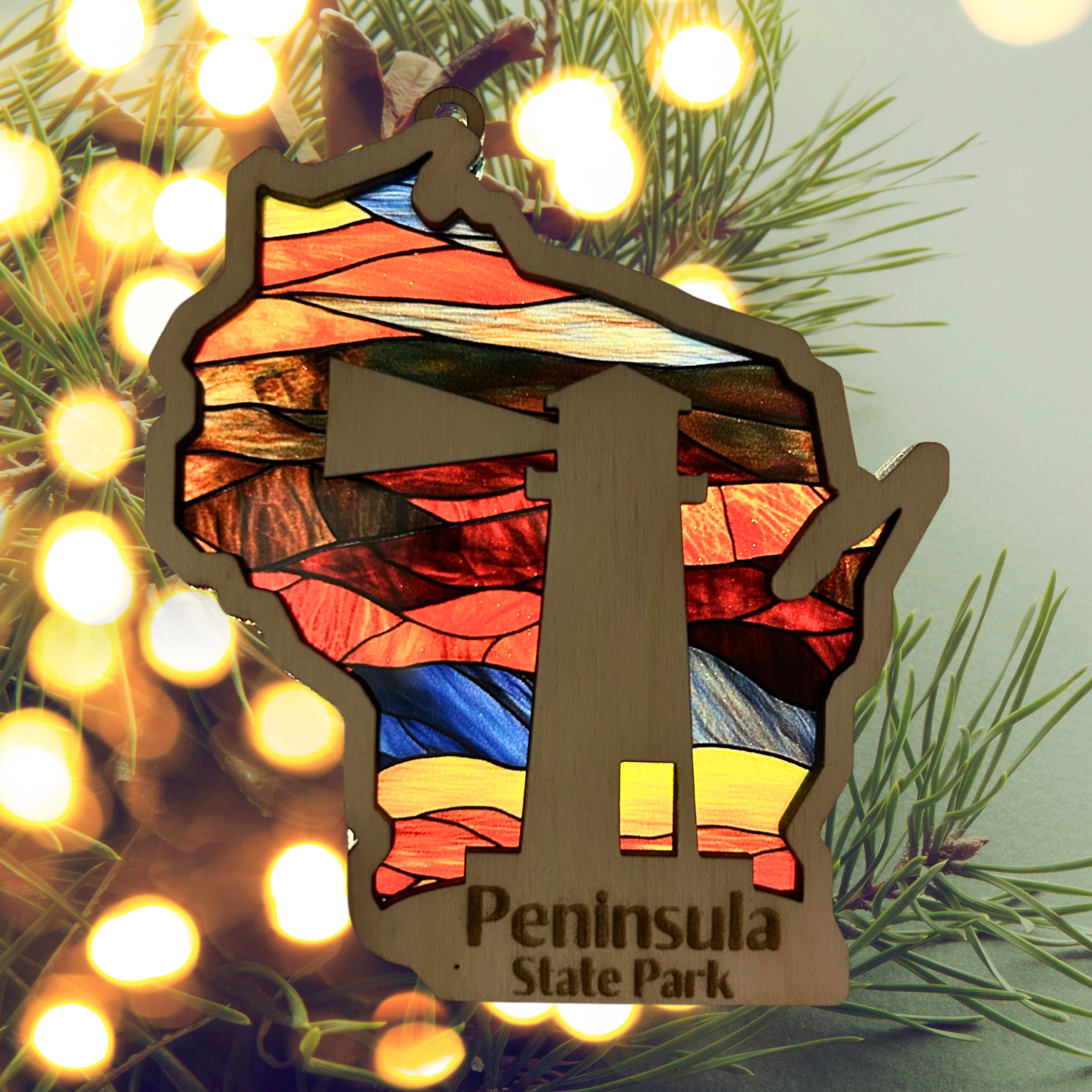Wood Wisconsin Shaped ornament with a stained glass look and a lighthouse.  Includes the text: Peninsula State Park. Photo Background: illuminated Christmas tree.
