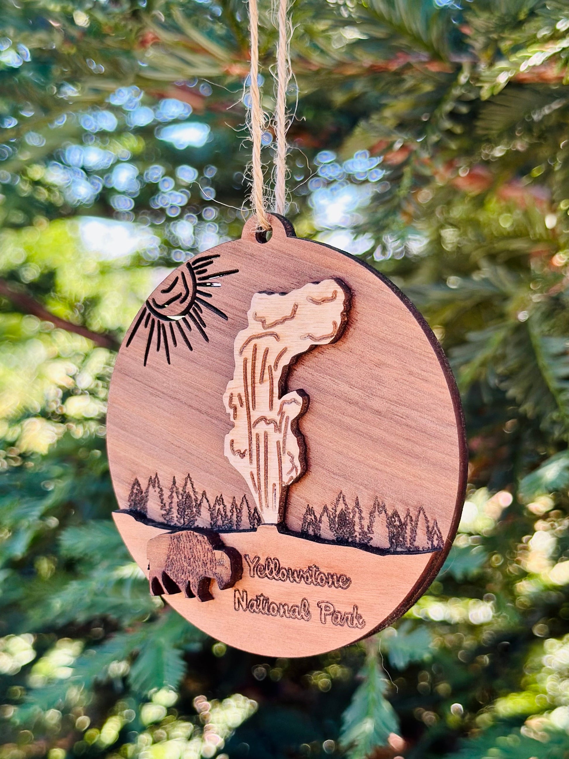 A round wooden hanging ornament engraved with Yellowstone National Park scenery, including a geyser, sun, trees, bison, and the park name, hanging from a string in front of blurred green foliage.