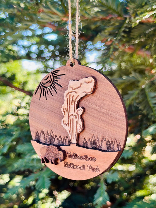 A round wooden hanging ornament engraved with Yellowstone National Park scenery, including a geyser, sun, trees, bison, and the park name, hanging from a string in front of blurred green foliage.