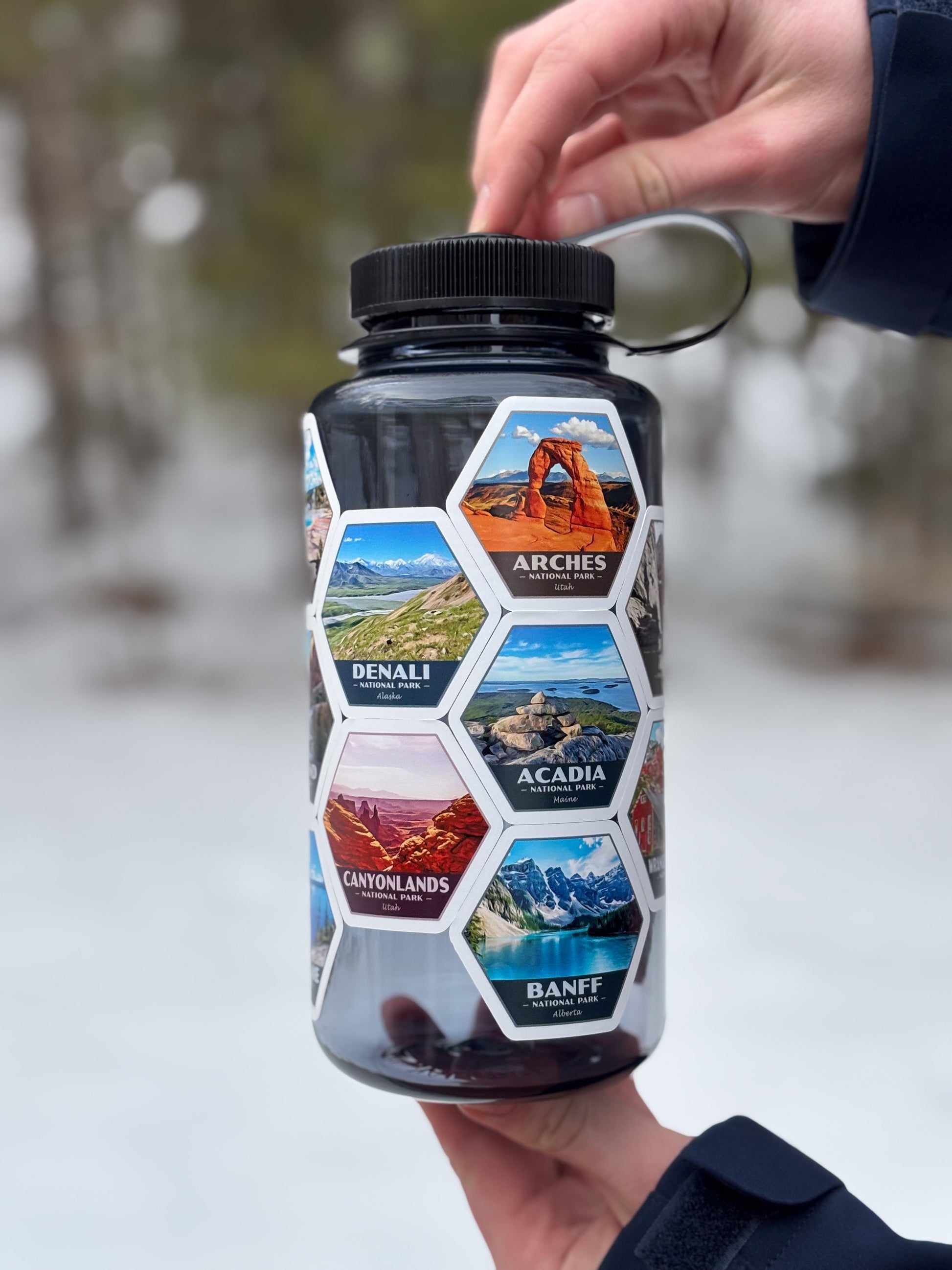 A person holds a clear plastic water bottle outdoors in a snowy setting. The bottle has a black screw cap and a metal loop handle, with a wraparound design made of hexagonal scenic photo tiles labeled Arches, Denali, Acadia, Canyonlands, and Banff.