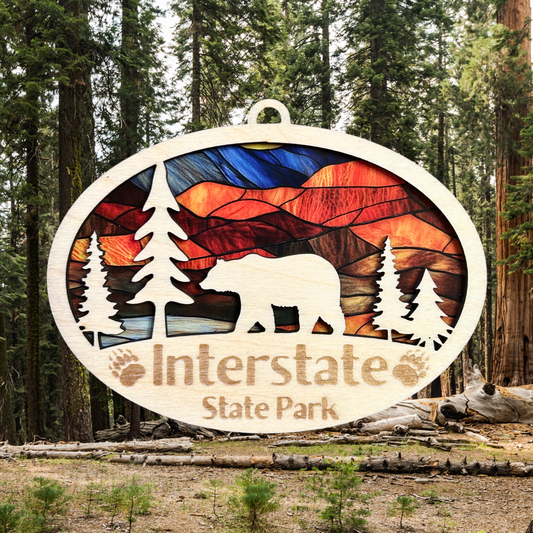 Oval shaped ornament with trees and bear and a stained glass appearance.  Includes the text “Interstate State Park” with engraved paw prints.  Photo Background: wooded forest.