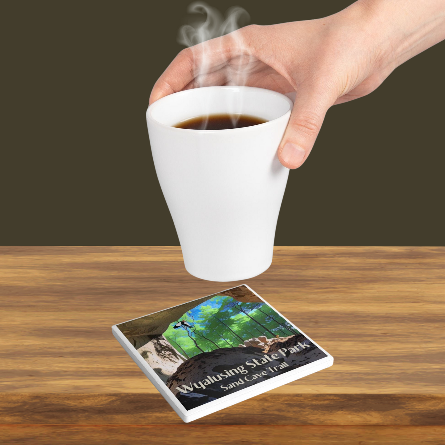 Ceramic coaster featuring a view from inside a sandstone cave looking out into a lush green forest with a woodpecker flying through the air.  Photo Background: coaster sitting on a wood counter surface with a person placing a hot beverage on it. 