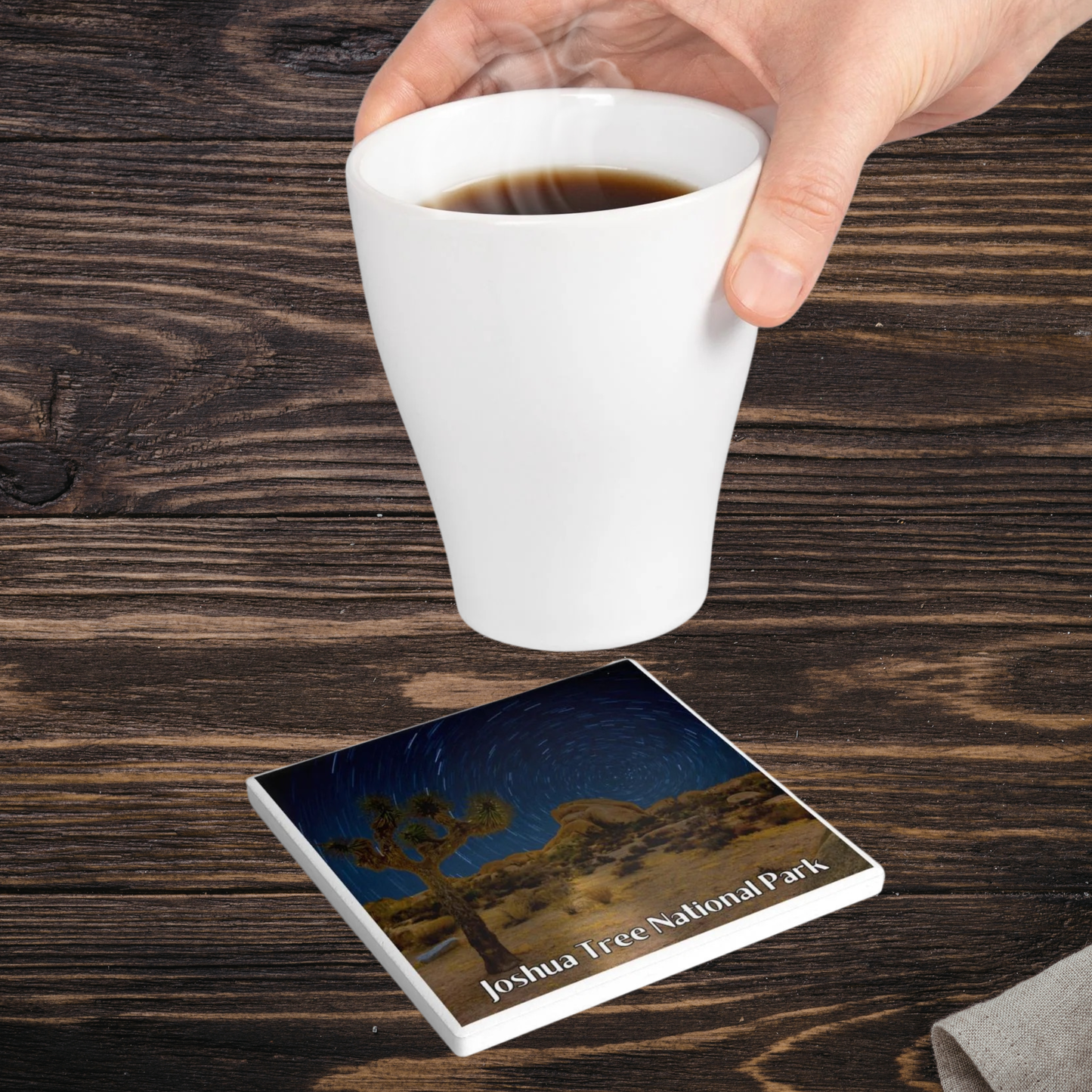Hand holding a white mug with coffee above a coaster featuring Joshua Tree National Park design on a wooden surface.