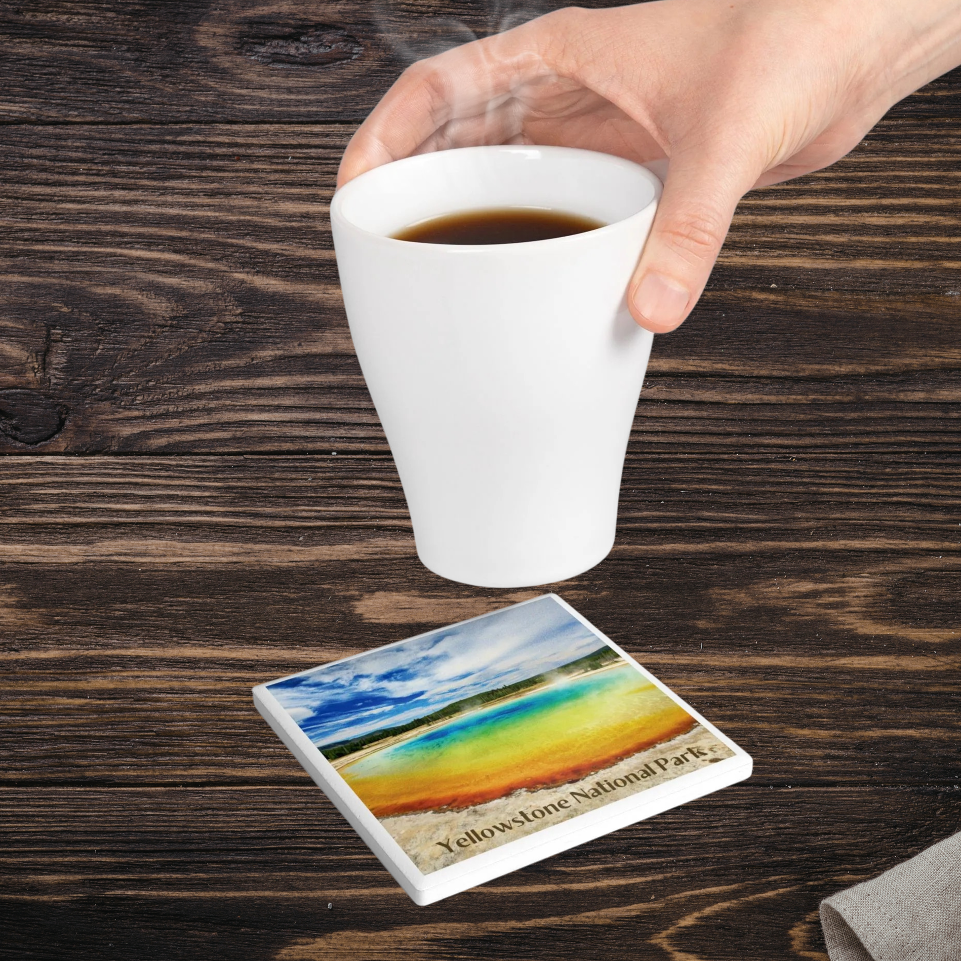 Hand holding a white mug with a scenic coaster featuring Yellowstone National Park on a wooden surface.