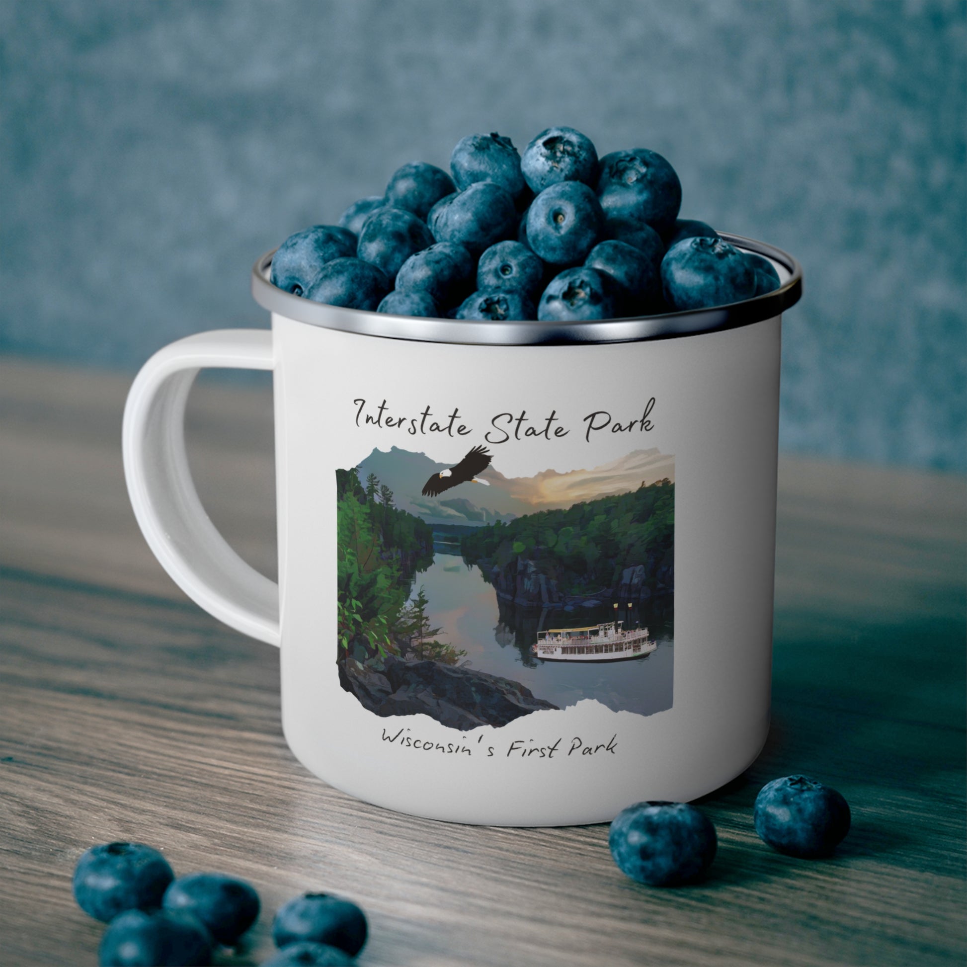 White enamel mug depicting the St Croix River at sunset with an eagle flying above and a paddle wheel boat on the river.  Photo background: mug sitting on a shelf full of blueberries.