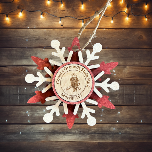 Handmade red layered snowflake with an eagle medallion and the text Council Grounds State Park Merrill, WI.  Photo background: Rustic wood with white Christmas lights.