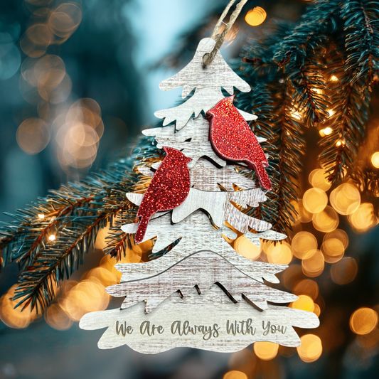 Two red glitter cardinals on a wood tree ornament with the text “we are always with you.” Photo background: closeup of lighted Christmas tree branch.