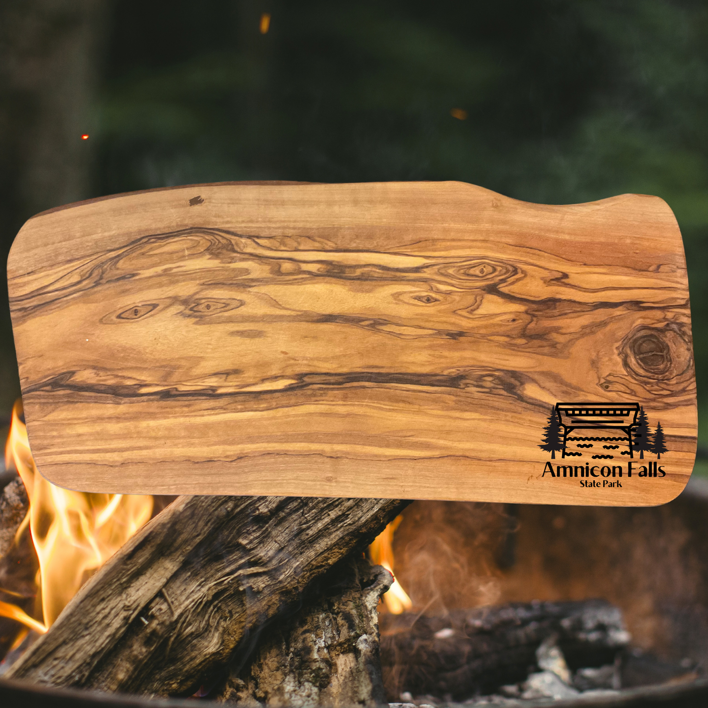 Olive wood serving board with a graphic depicting a covered bridge over a stream with trees and the text Amnicon Falls State Park.  Photo background: campfire at night.