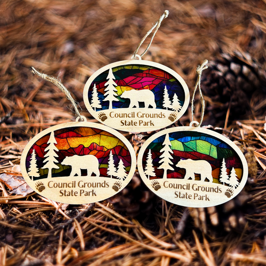 Three handmade oval wood ornaments featuring a bear walking amongst trees on a stained glass appearance background.  Photo background: pine needles and pinecones. 