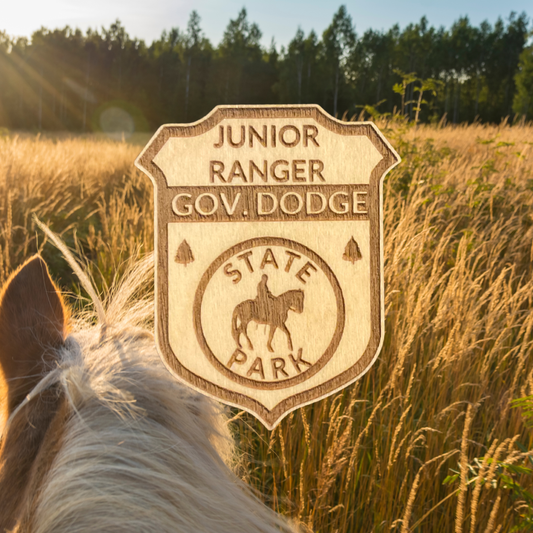Laser engraved wood badge featuring the text: “Junior Ranger Gov Dodge State Park” with the icon of a person riding a horse.  Photo Background: Horse walking through a golden field of tall grass.