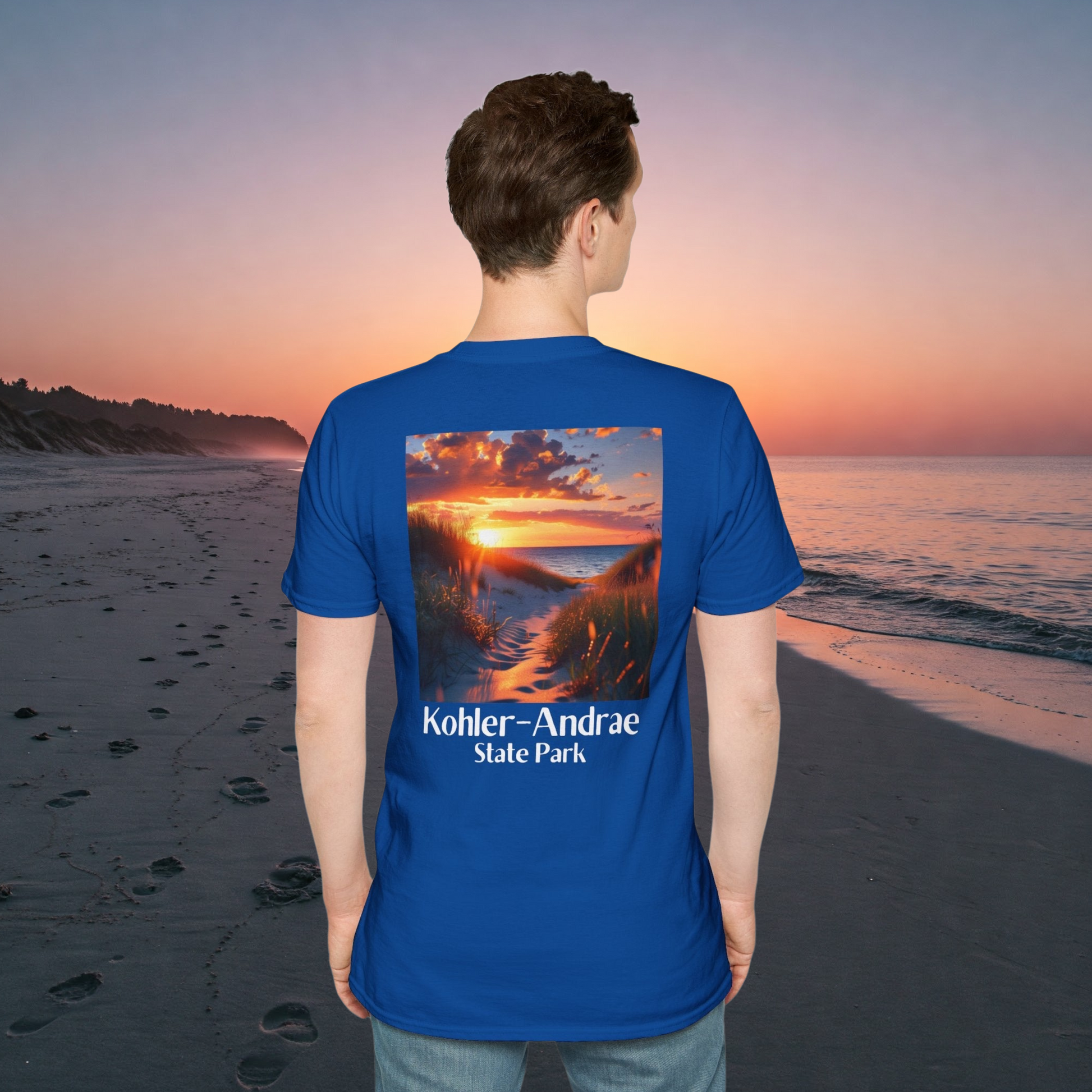 Male Model standing on a sandy beach at sunrise wearing a royal t-shirt featuring a back design of looking through the sand dunes at a colorful sunrise over Lake Michigan and the text: Kohler-Andrae State Park. 