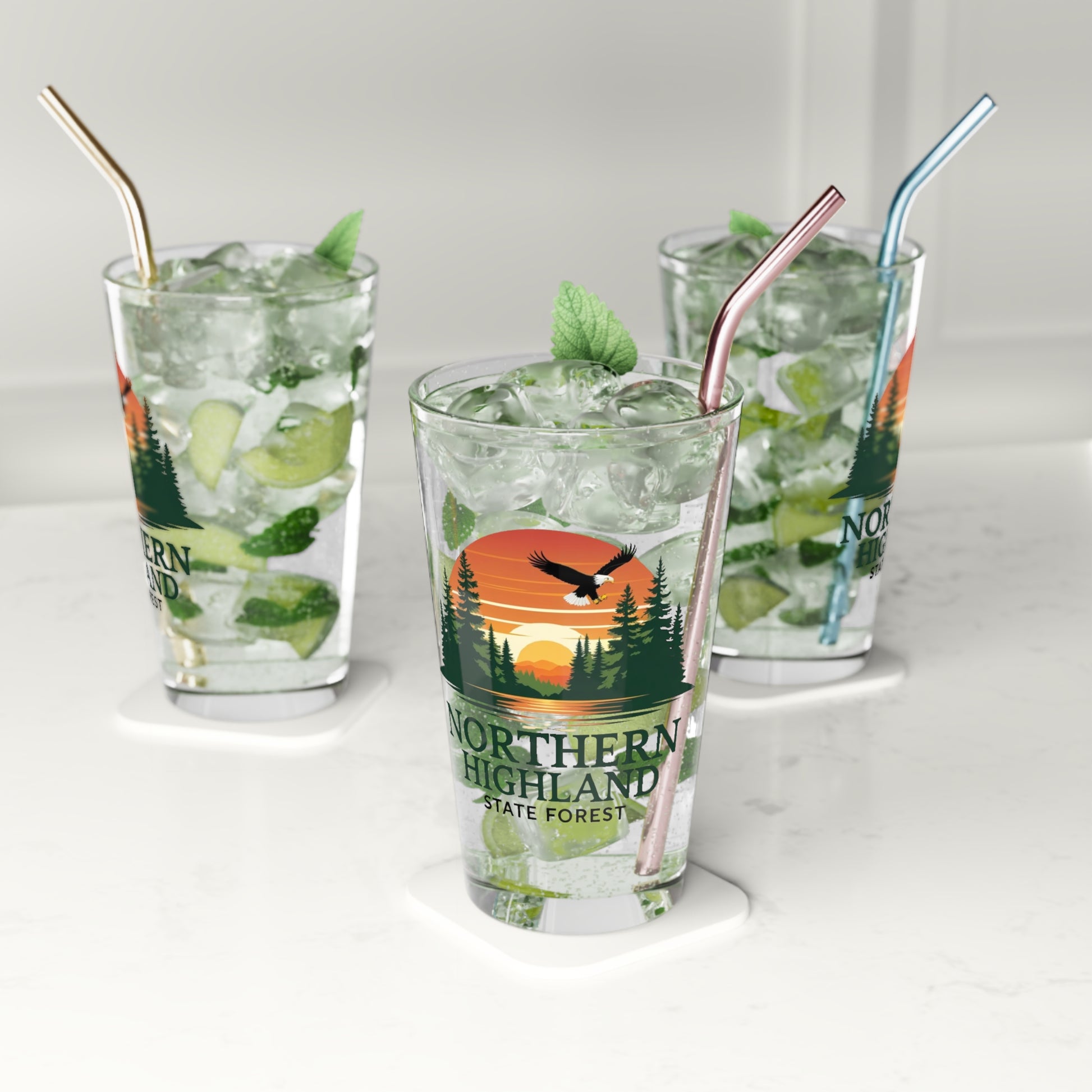 16 oz glass pint featuring a colorful design of a sunset over a lake surrounded by pine trees and a Bald Eagle Soaring overhead, with the text Northern Highland State Forest.  Photo background: three glasses sitting on the counter filled with water and lime wedges. 