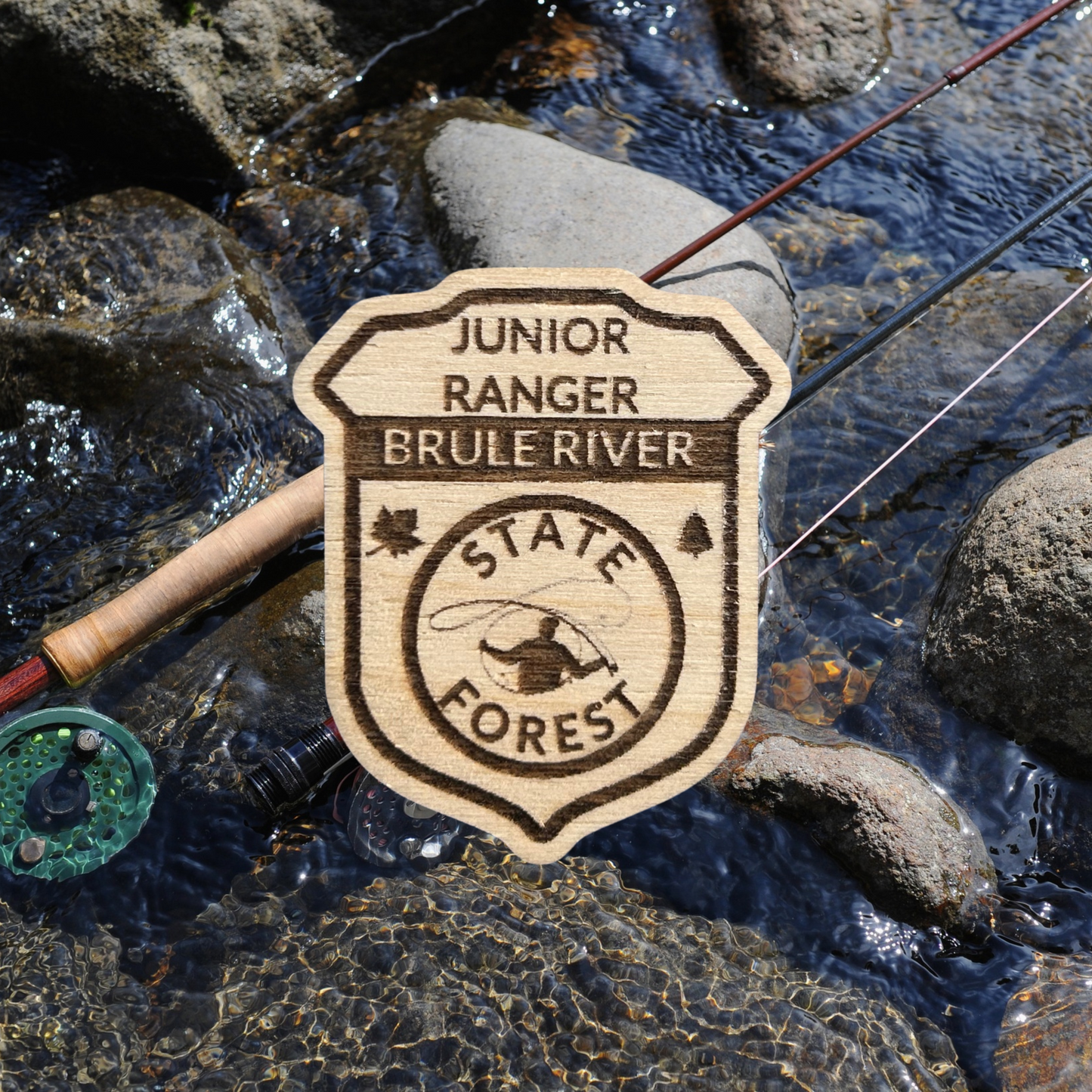 Wood Jr Ranger badge with the text: “Junior Ranger Brule River State Forest” and an icon of a fly fisherman casting.  Photo background: two fly rods laying on the rocks in a stream.