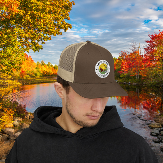 Male model wearing a Brown and Khaki Colored ball cap with embroidered logo of an angler catching a fish and the text Brule River State Forest. Photo background is of trees in fall colored foliage on the banks of a river. 