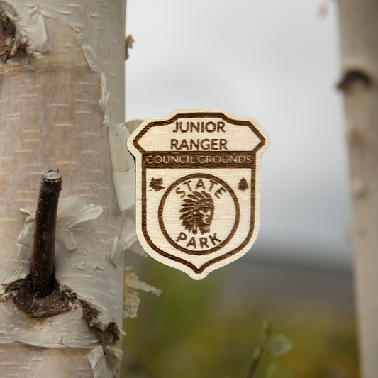 Wood badge shaped JR Ranger Badge with the text “Junior Ranger, Council Grounds State Park” with a Indian Head symbol.  Photo background: white birch trees.