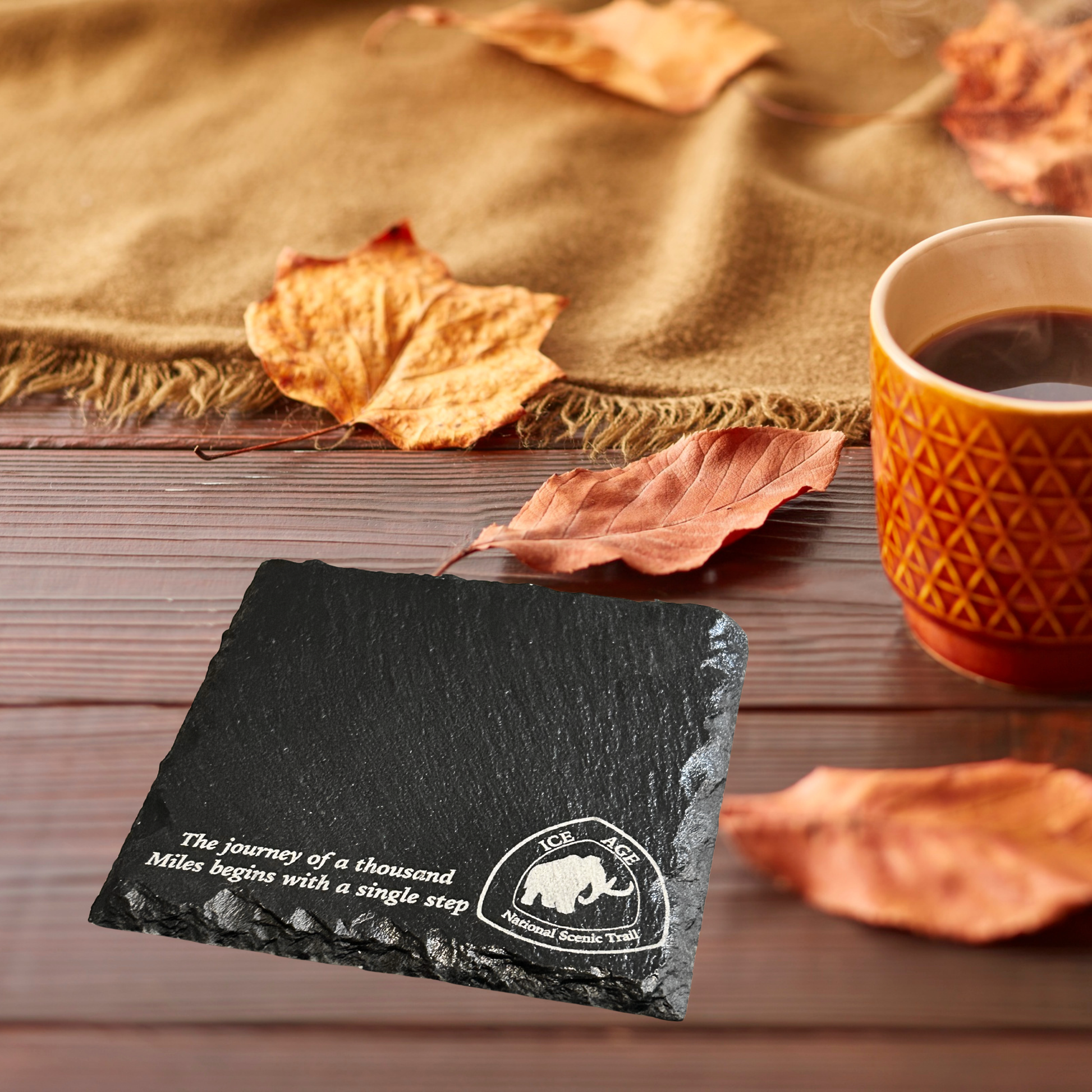 Slate coaster with text and logo on a wooden surface with a cup of coffee and autumn leaves.