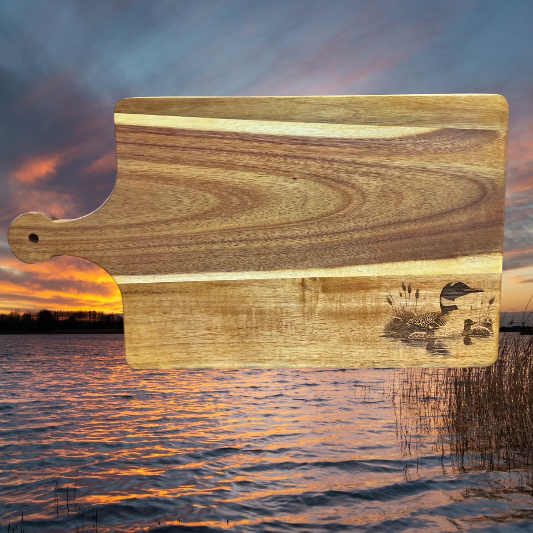 Acacia wood cutting board with a loon and two chicks engraved on it.  Lake sunset background. 
