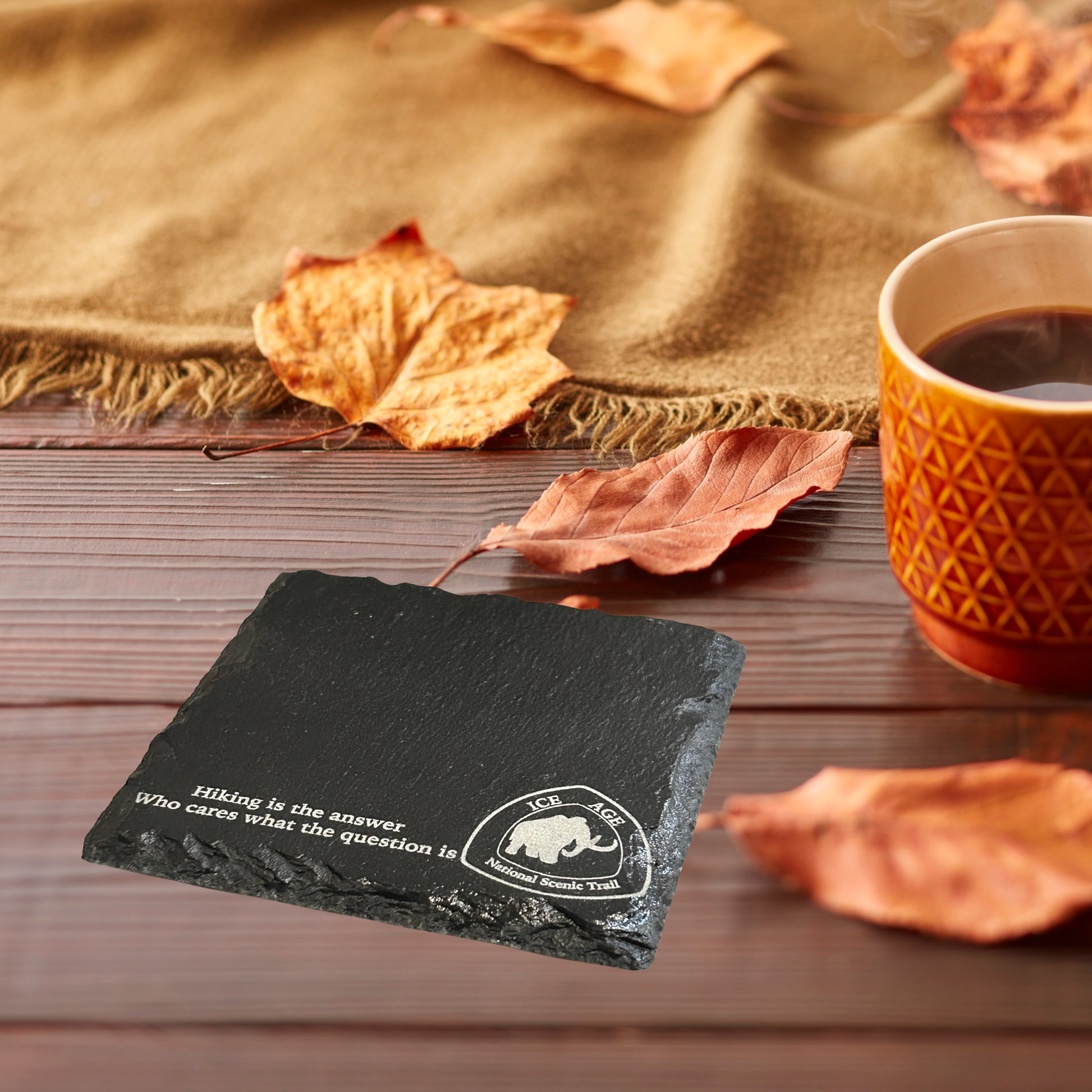 Slate coaster with text and logo on a wooden surface with a cup of coffee and autumn leaves.