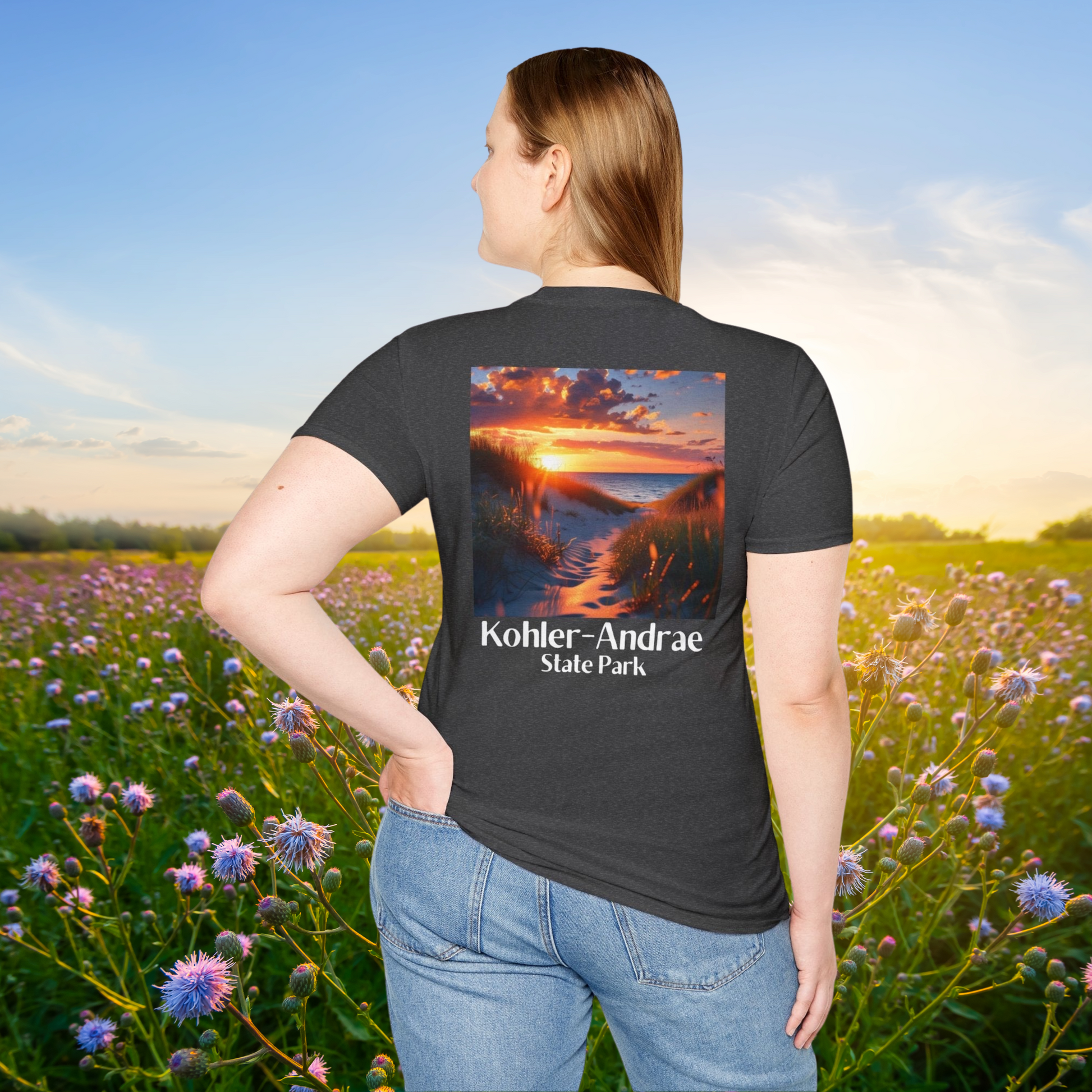 Female model standing in a field of wild flowers wearing a dark heather t-shirt featuring a back design of looking through the sand dunes at a colorful sunrise over Lake Michigan and the text: Kohler-Andrae State Park.