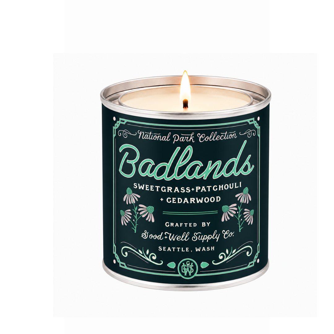 A lit metal tin candle from the National Park Collection, specifically the Badlands scent, featuring a dark teal label with white and mint green typography and botanical illustrations.