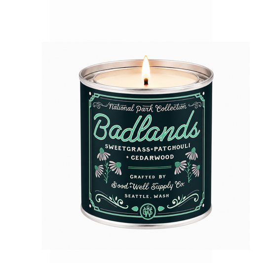 A lit metal tin candle from the National Park Collection, specifically the Badlands scent, featuring a dark teal label with white and mint green typography and botanical illustrations.