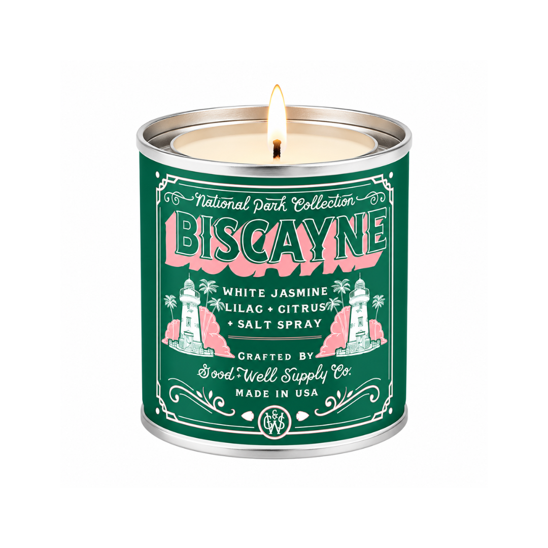 A lit scented candle in a cylindrical metal tin with a dark green label reading 'National Park Collection Biscayne' and 'White Jasmine, Lilac, Citrus, + Salt Spray.' The tin has silver metal rims and a visible flame at the top, photographed on a plain white background.