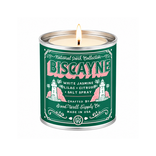 A lit scented candle in a cylindrical metal tin with a dark green label reading 'National Park Collection Biscayne' and 'White Jasmine, Lilac, Citrus, + Salt Spray.' The tin has silver metal rims and a visible flame at the top, photographed on a plain white background.