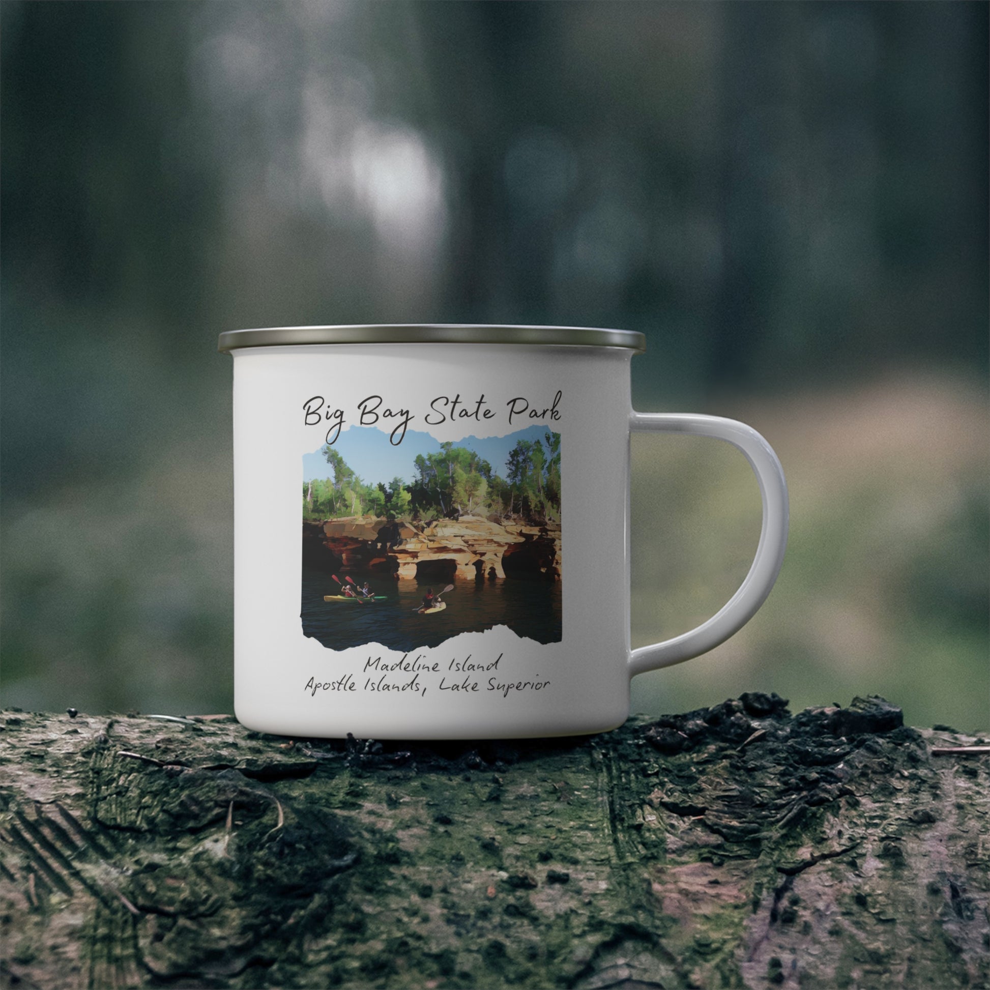 White enamel mug with picture depicting kayakers in front of sea caves and the text “Big Bay State Park - Madeline Island, Apostle Islands, Lake Superior.  Photo background: mug sitting on log in woods.